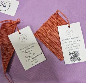 A photo of two orange crochet swatches created by Sammi during the 2025 SoCal Fiber Fest. Each swatch has a Yarn Over Hook branded tag attached. One tag lists the maker’s name, yarn type (‘fingering wool/nylon blend’), and hook size (‘3 mm’). The other tag invites viewers to work with Yarn Over Hook and includes a QR code. Everything is displayed on a lavender-purple background.