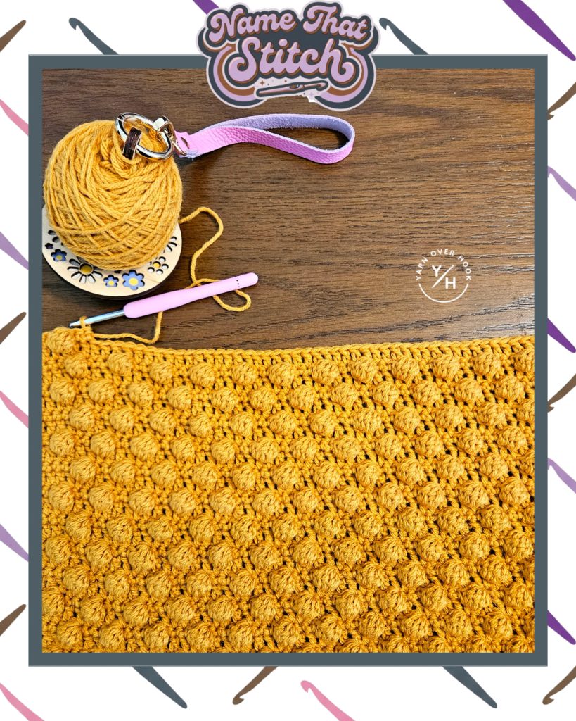 Close-up of a crocheted fabric featuring a repeating textured stitch pattern in mustard yellow yarn, shown alongside a yarn ball and crochet hook, featured as part of the Name That Stitch daily crochet puzzle.