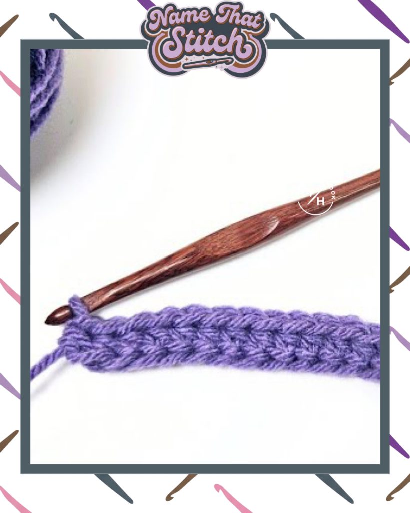 Close-up of a crochet work-in-progress showing a short row of textured stitches in purple yarn, with a wooden crochet hook and yarn ball, featured as part of the Name That Stitch daily crochet puzzle. View more at NameThatStitch.com