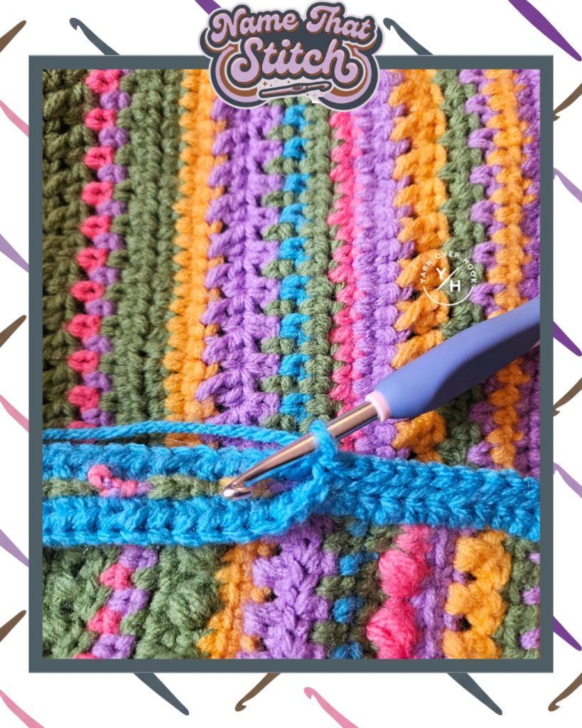 Close-up of a colorful crocheted fabric with vertical rows in green, purple, orange, and pink yarn, showing a blue row in progress with a crochet hook, featured as part of the Name That Stitch daily crochet puzzle. View more at NameThatStitch.com