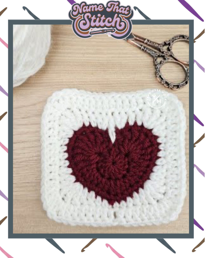 Square crochet swatch featuring a textured red heart centered inside a white border, photographed on a light wood surface with a ball of white yarn and small vintage-style scissors nearby for a Valentine’s Day Name That Stitch puzzle. View more at NameThatStitch.com.