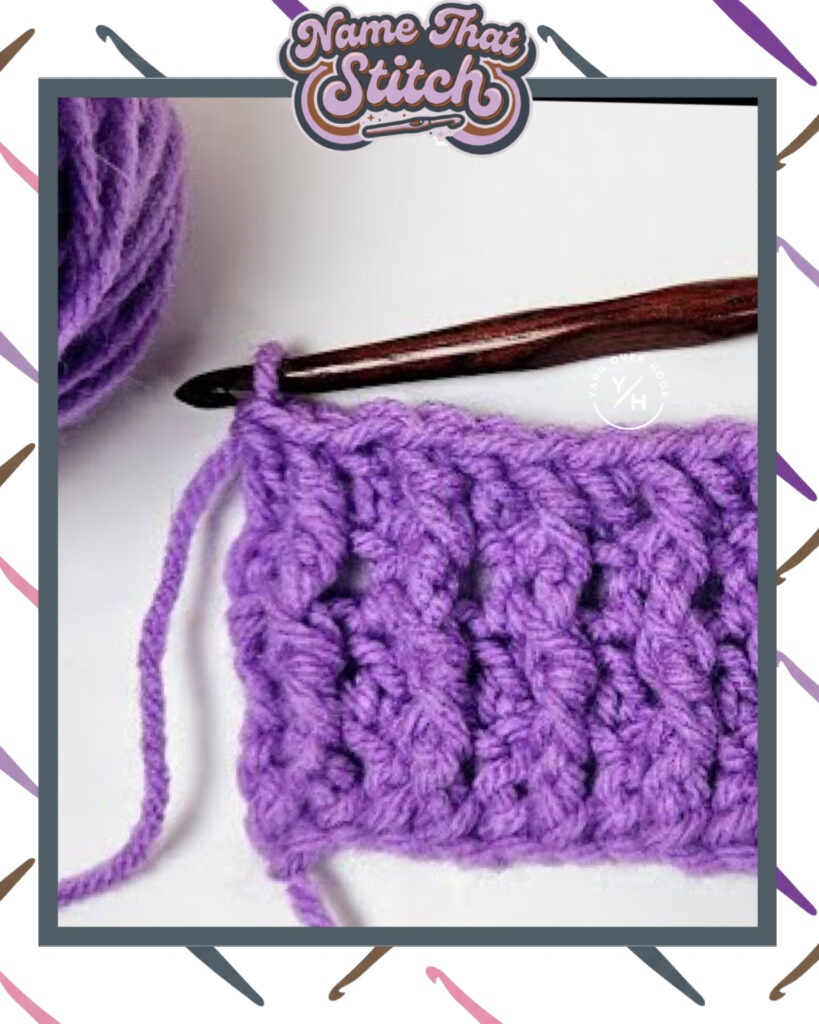 Close-up of a crocheted fabric displaying a repeating stitch pattern worked in purple yarn, with a wooden crochet hook and featured as part of the Name That Stitch daily crochet puzzle. View more at NameThatStitch.com