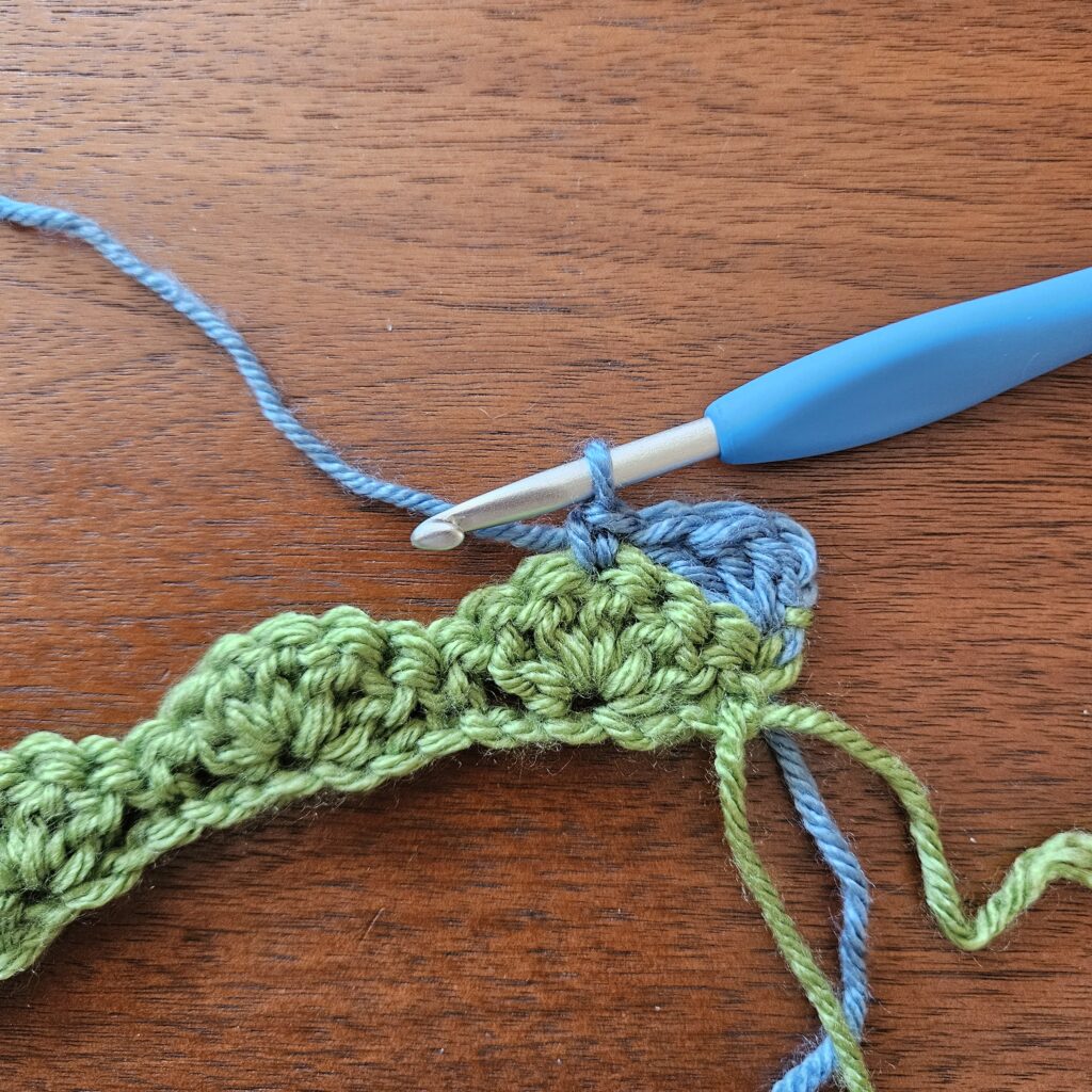 Close-up of a crochet hook working blue yarn into a row of textured green crochet stitches on a wooden table and featured as part of the Name That Stitch daily crochet puzzle. View more at NameThatStitch.com