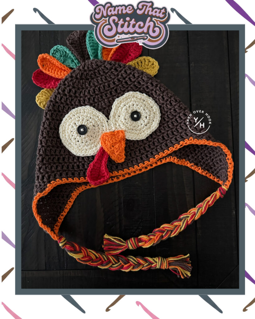 Name That Stitch game card showing a crocheted turkey hat in brown, cream, orange, red, and gold with large eyes, a beak, colorful feathers, and braided ties with Yarn Over Hook branding. View more at NameThatStitch.com