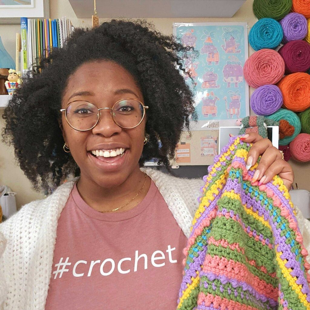 Smiling crochet instructor, Sammi Jai from Yarn Over Hook, holding a colorful striped crochet project in a bright yarn-filled studio