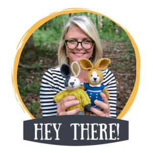 Woman (Elise Rose) with glasses smiling outdoors while holding two crocheted bunny dolls, framed by a yellow circle with the text “Hey There!” below.