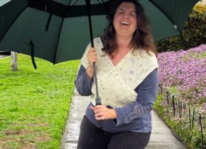 Alexis Kavros smiling outdoors under a dark green umbrella while wearing a crocheted cream top, with purple flowers in the background.