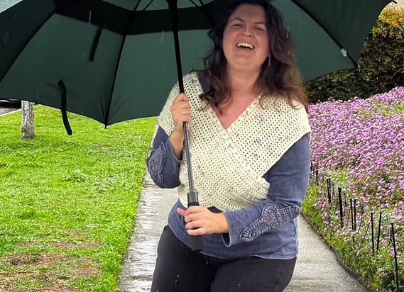 Alexis Kavros smiling outdoors under a dark green umbrella while wearing a crocheted cream top, with purple flowers in the background.