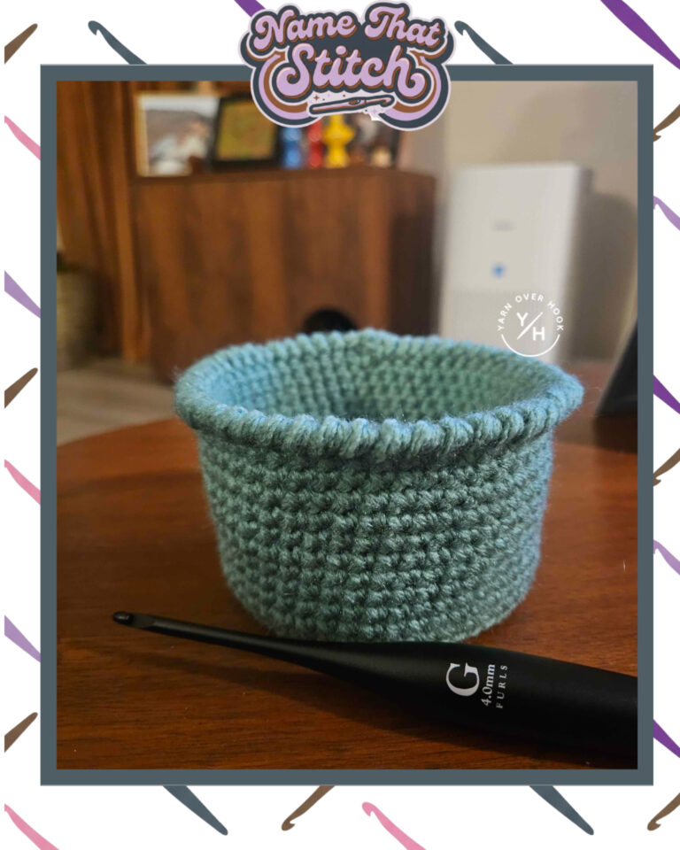 Close-up of a small blue crocheted basket or container displaying a dense repeating stitch pattern beside a black crochet hook on a wooden table, featured as part of the Name That Stitch daily crochet puzzle. View more at NameThatStitch.com