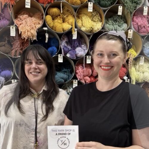 Two women standing in front of a wall of colorful yarn cones, holding a Yarn Over Hook sign that reads, “This yarn shop is a friend of Yarn Over Hook.”