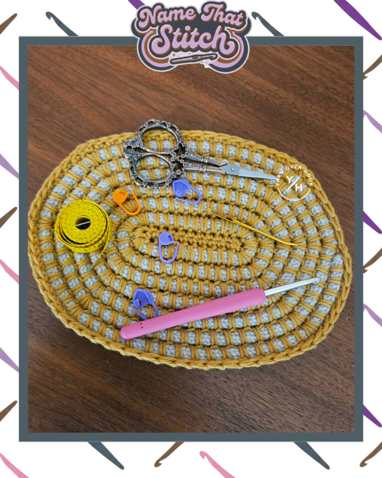 Close-up of an oval crocheted mat in mustard yellow and gray holding scissors, stitch markers, a tape measure, and crochet hooks, displaying a repeating openwork stitch pattern and featured as part of the Name That Stitch daily crochet puzzle. View more at NameThatStitch.com