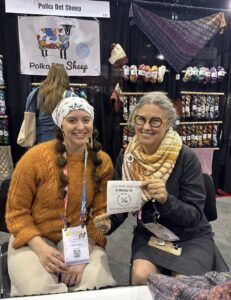 Polka Dot Sheep staff members smiling at a fiber arts booth, surrounded by colorful yarn skeins, knit samples, and a Polka Dot Sheep banner.