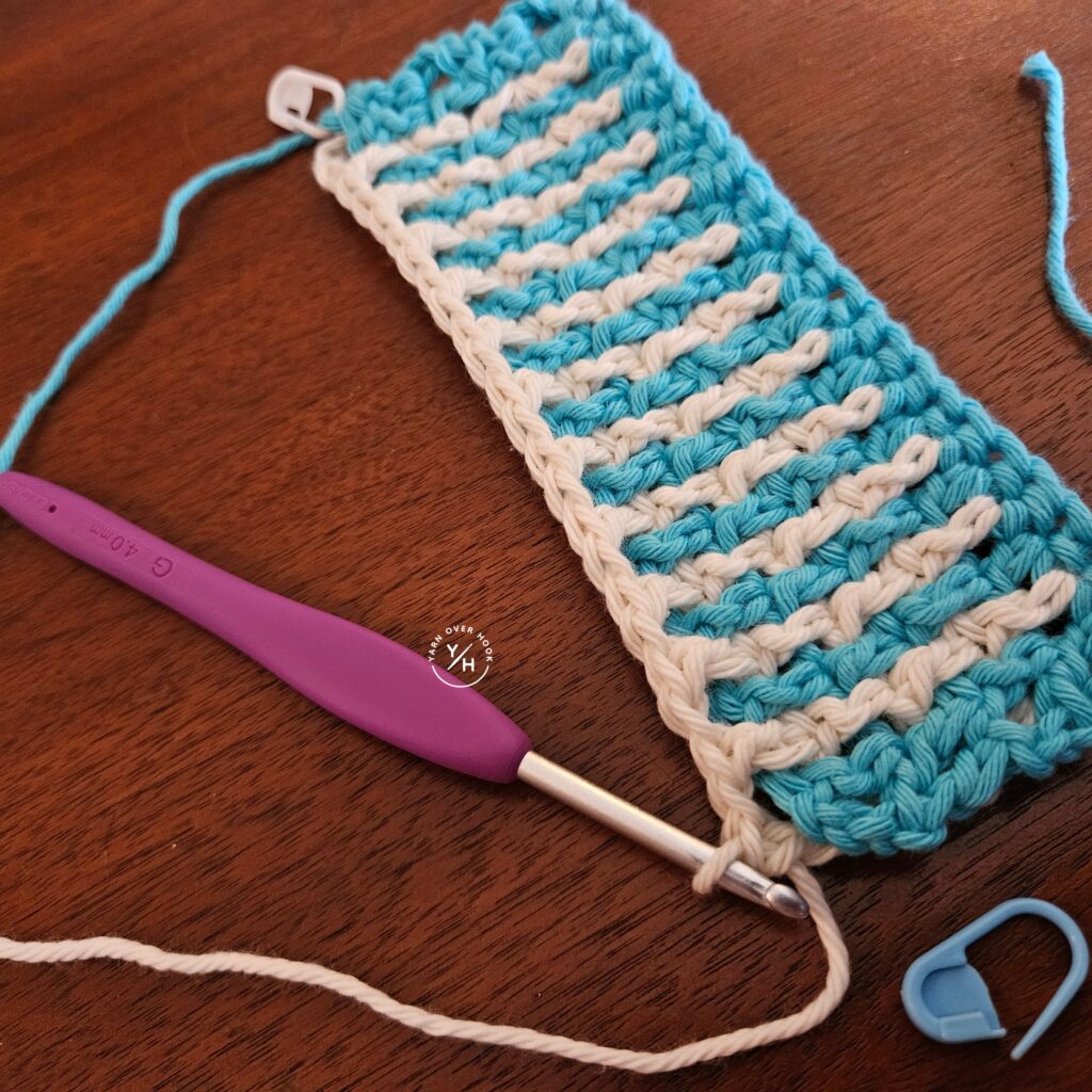 Clover Amour crochet hook with a purple handle resting beside a turquoise and white crochet stitch swatch in progress.