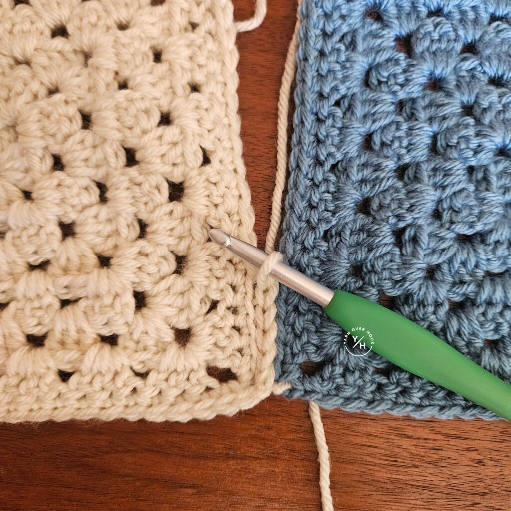 Clover ergonomic crochet hook with a green handle joining cream and blue granny stitch crochet squares on a wooden table.