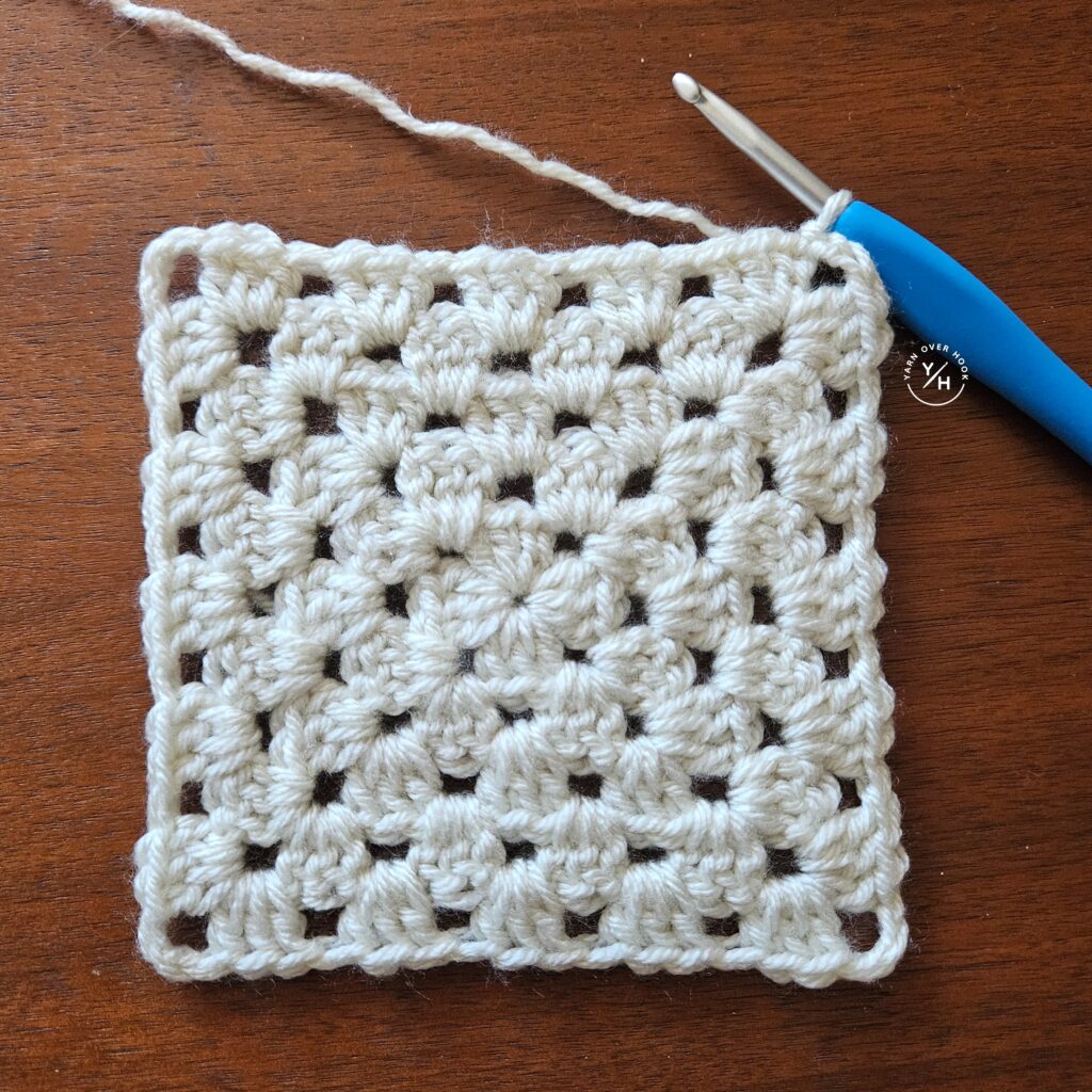Clover ergonomic crochet hook with a blue handle resting beside a white granny stitch crochet swatch on a wooden table.