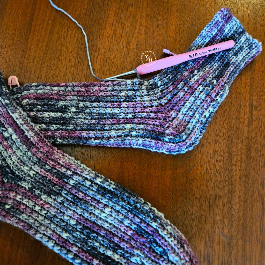 Tulip ergonomic crochet hook with a pink handle resting on a variegated purple, blue, and cream crochet project in progress.
